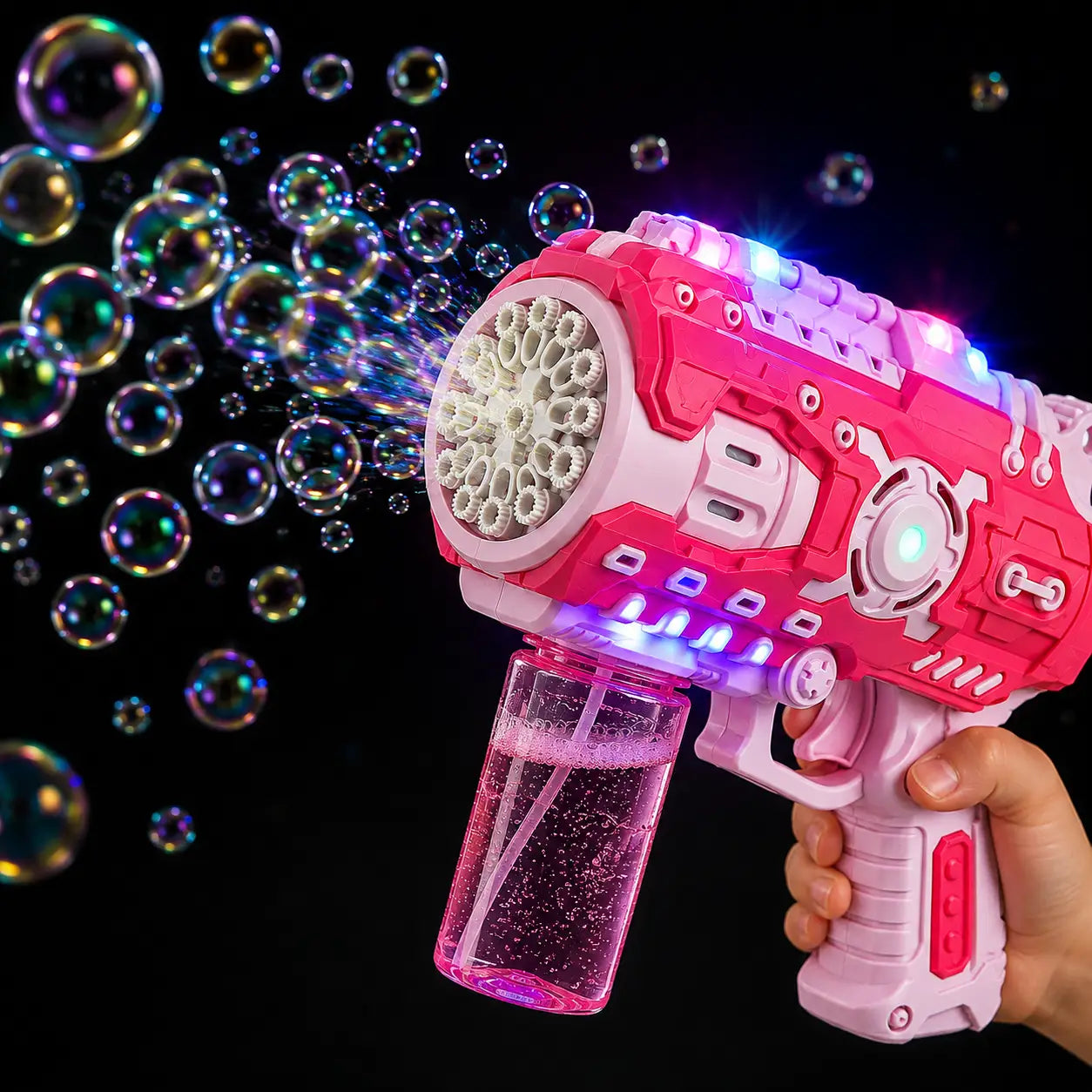 A pink and white toy bubble gun with glowing LED lights and a clear liquid reservoir, shown with bubbles spraying out against a black background. A hand is holding the toy.