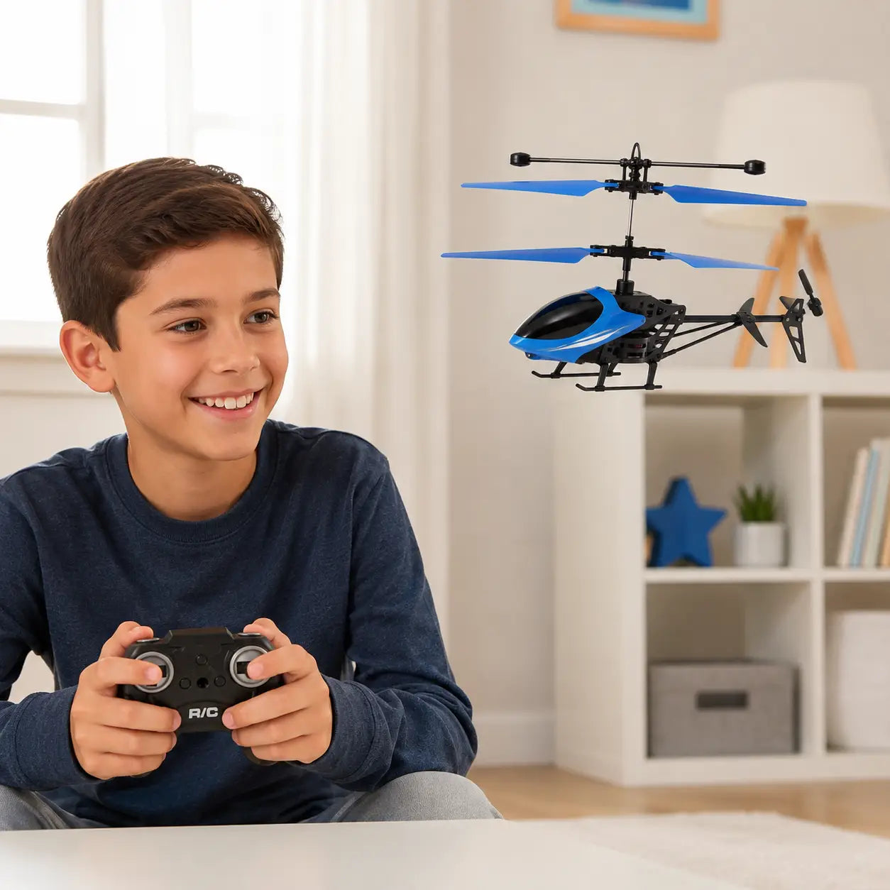 A young boy wearing a navy blue long-sleeve shirt smiles while holding a black remote control in his hands. A blue and black remote-controlled toy helicopter floats in the air in front of him, set against a bright, modern room background with white walls and shelves.